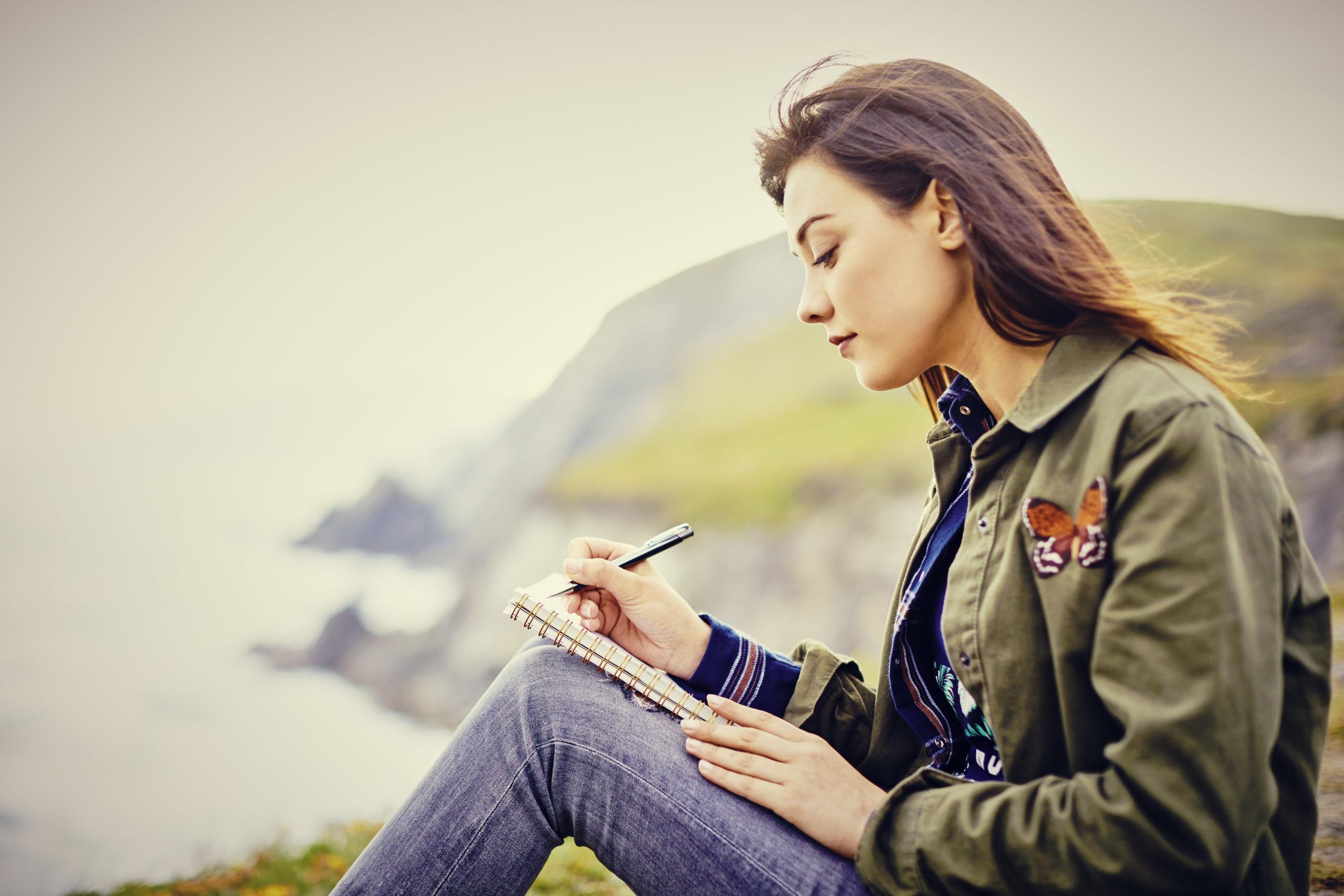 Side view of young woman writing in diary while sitting on mountain. Beautiful female tourist spending leisure time in nature. She is wearing casuals.