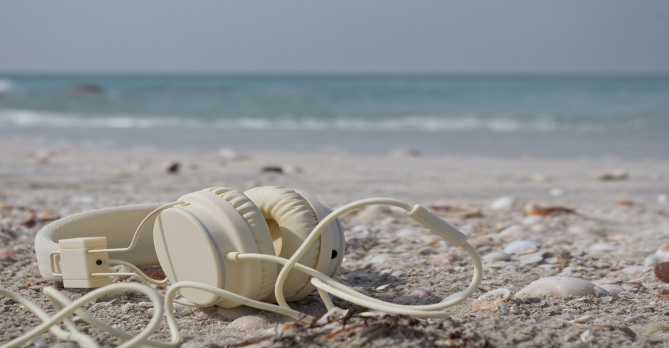 Valkoiset kuulokkeet hiekkarannalla, jolla myös simpukankuoria, taustalla aaltoilee meri.
