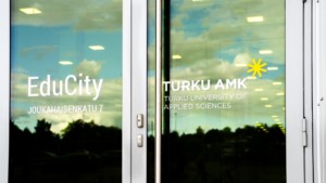 EduCityn ulko-ovi, johon heijastuu rakennuksia ja taivasta. Ovessa on Turun ammattikorkeakoulun logo.