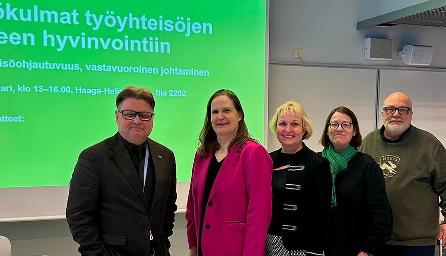 Viisi henkilöä seisovan tekstiesityksen edessä.